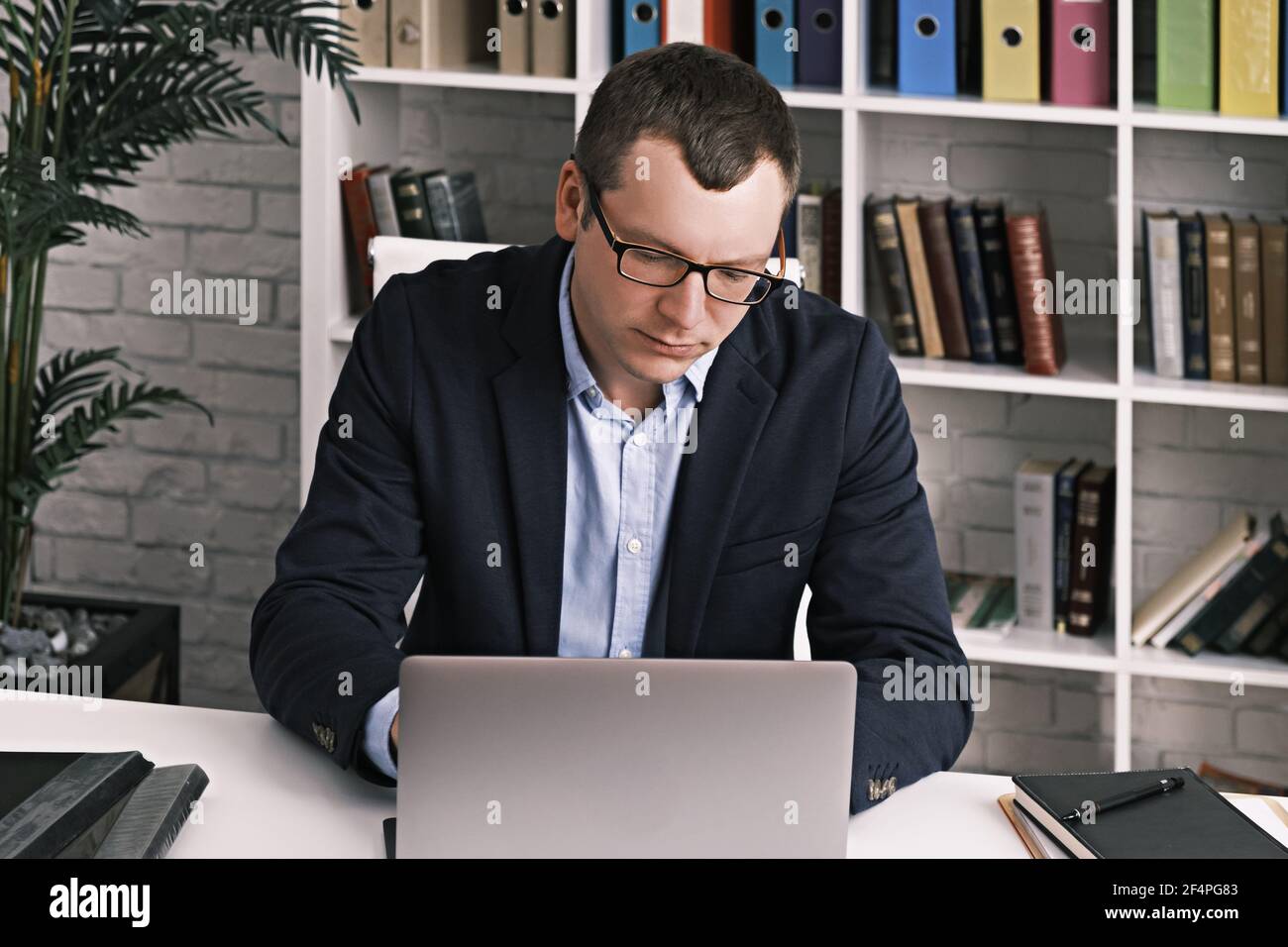 Un uomo serio messo a fuoco in occhiali e un vestito di affari stampa qualcosa usando un laptop seduto in ufficio leggero contro un rack con le cartelle. Lavoro in ufficio, Foto Stock