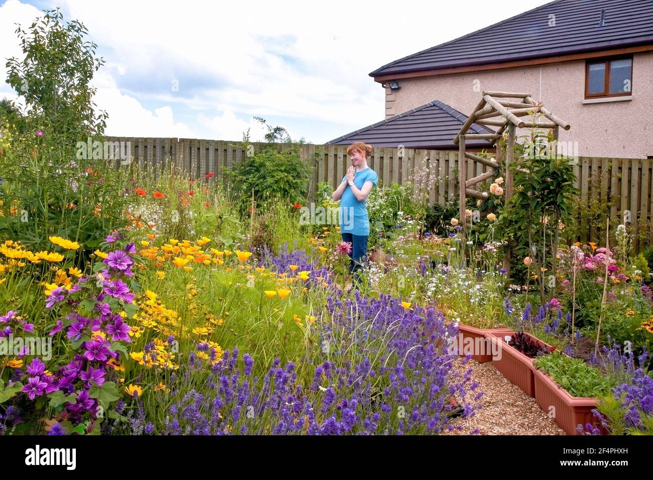 Vista del grazioso giardino in stile cottage sensoriale pieno di fiori estivi e giovane donna in piedi ammirando in gratitudine con le mani in posizione di preghiera. Foto Stock