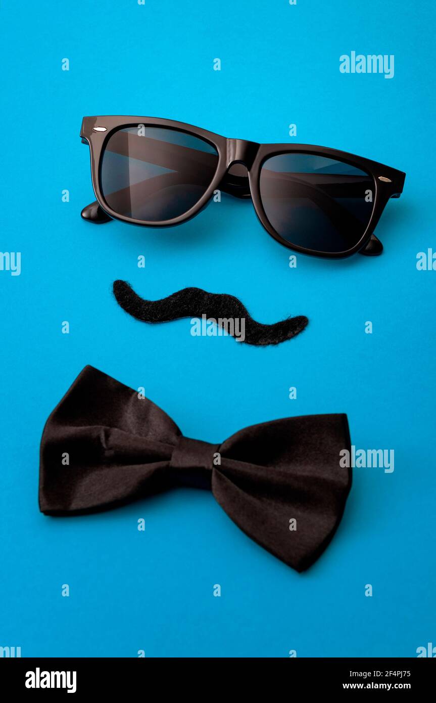 Il concetto di moda del giorno del padre e dell'hipster maschile con l'immagine minimalista di un paio di occhiali da sole quadrati, bowtie nere e un falso baffi su blu colorato b Foto Stock