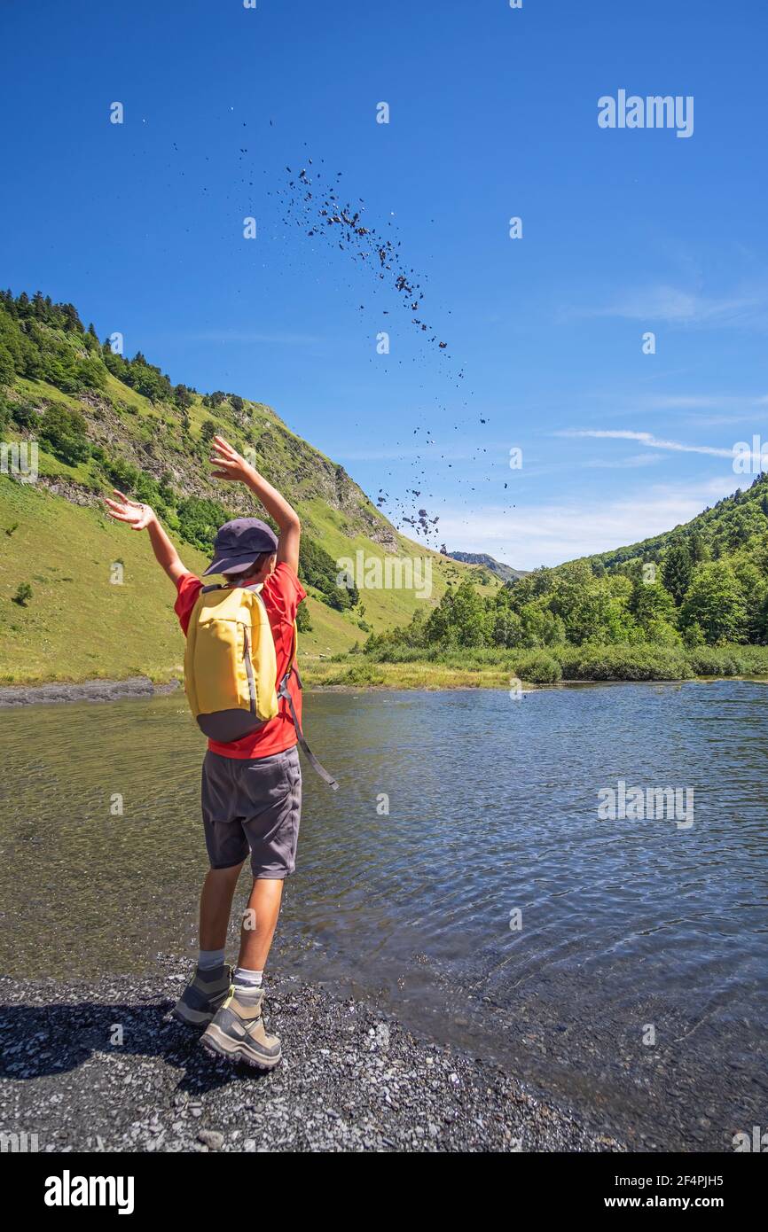 Boy explorer vestito con una T-shirt arancione, cappuccio e zaino giallo, getta pietre nell'aria in un lago di montagna Foto Stock