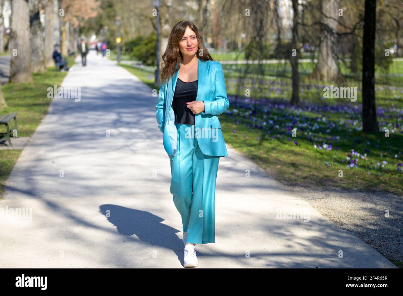 Elegante e attraente donna di mezza età con pantaloni turchesi che si adattano lungo un sentiero in un parco boscoso in primavera che guarda fuori a lato in un intero len Foto Stock