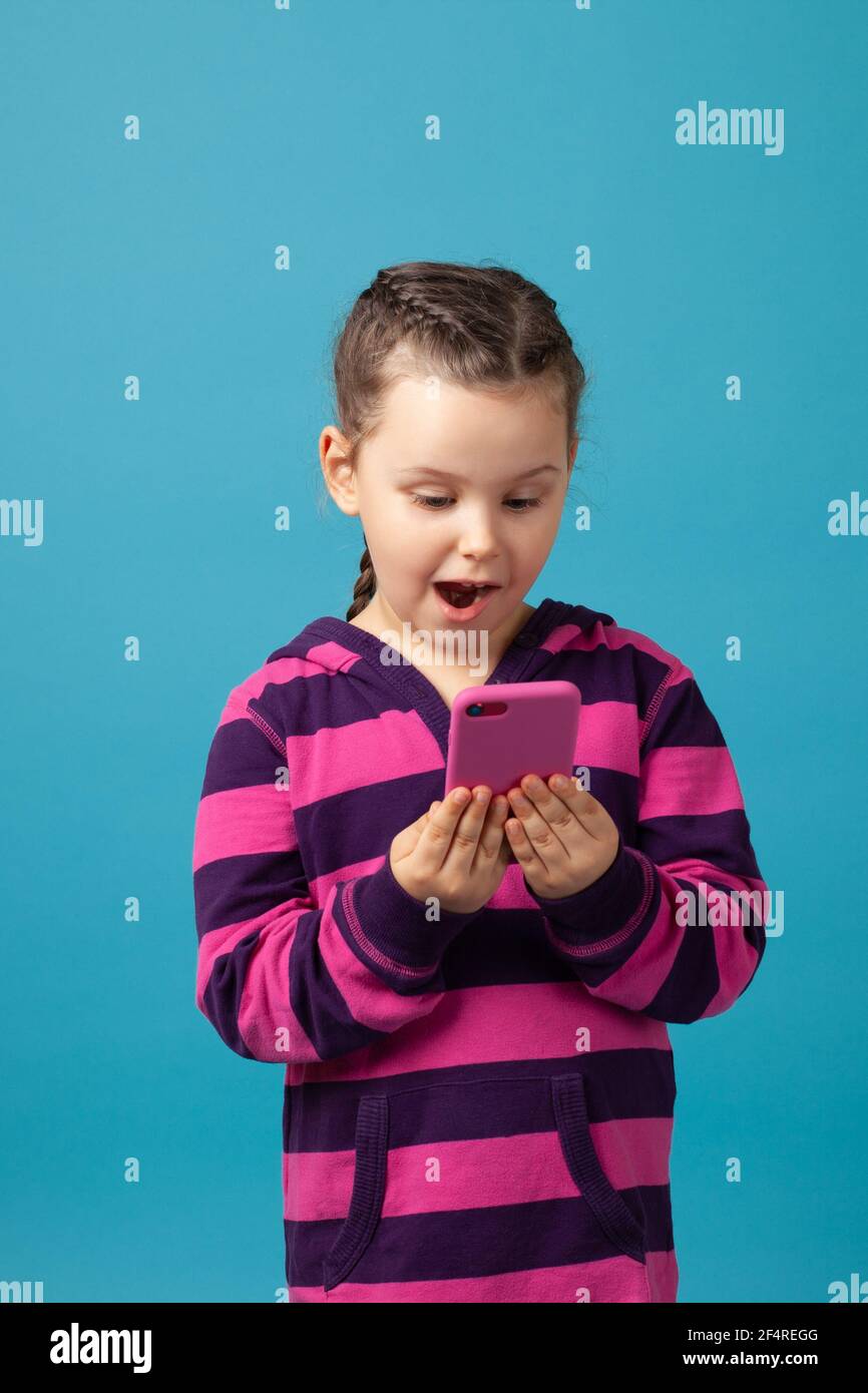 bambina con pigtail entusiasticamente e felicemente guardando il telefono in mano, comunicando con parenti o amici, isolato su sfondo blu Foto Stock