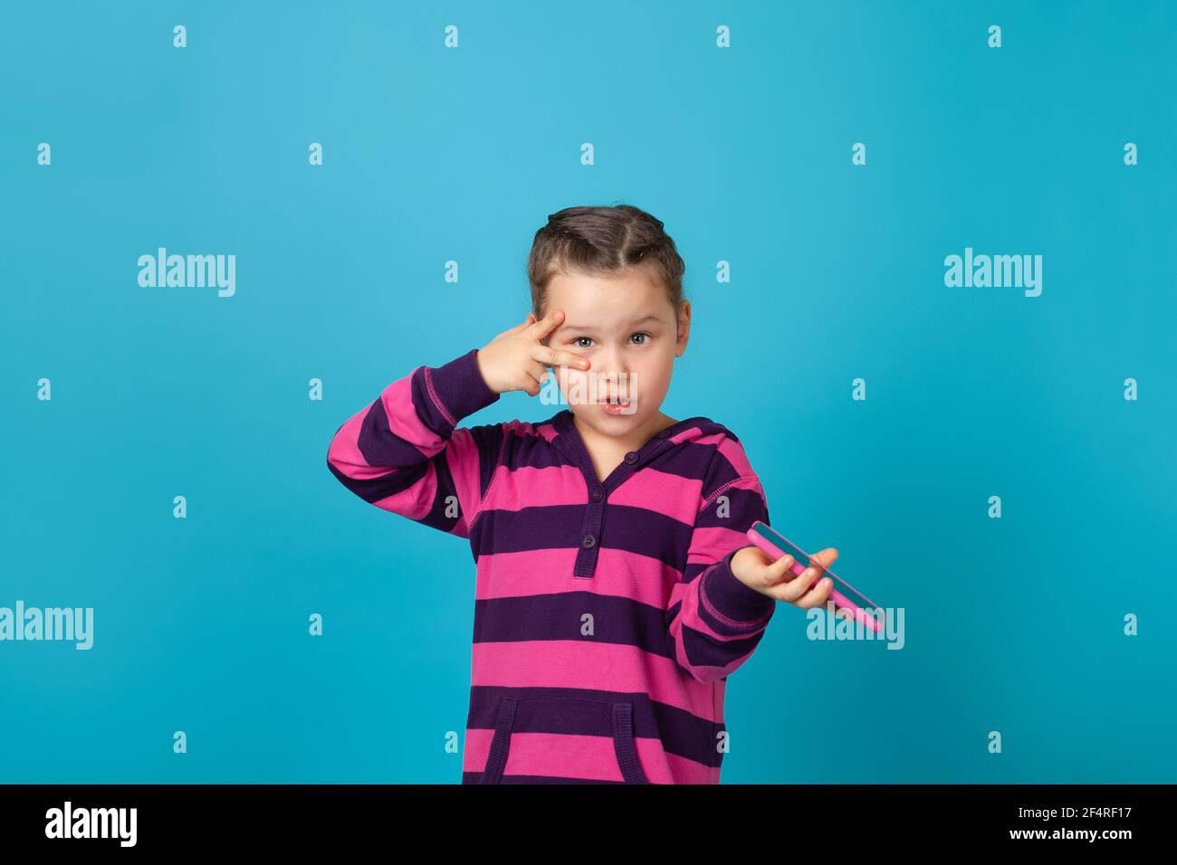 ritratto dinamico di ragazza con pigtail guardando la macchina fotografica, tenendo il telefono in mano e mostrando il segno della vittoria vicino all'occhio, isolato su uno sfondo blu Foto Stock