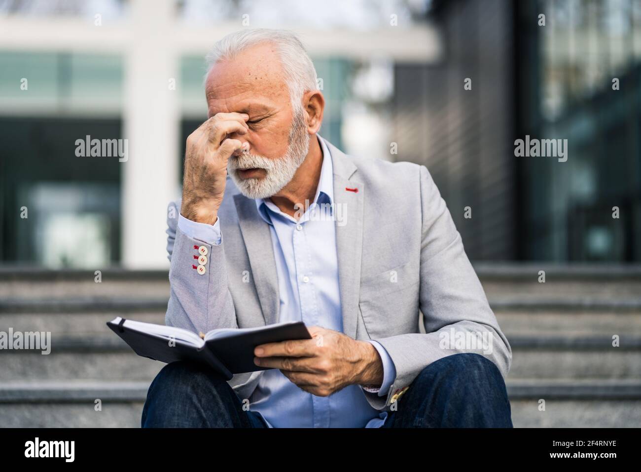 L'uomo d'affari anziano è stanco di lavoro. È seduto di fronte alla costruzione dell'azienda e ha mal di testa. Foto Stock
