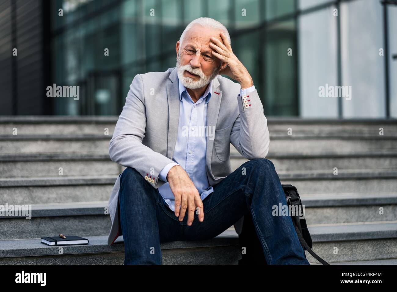 L'uomo d'affari anziano è stanco di lavoro. È seduto di fronte alla costruzione dell'azienda e ha mal di testa. Foto Stock