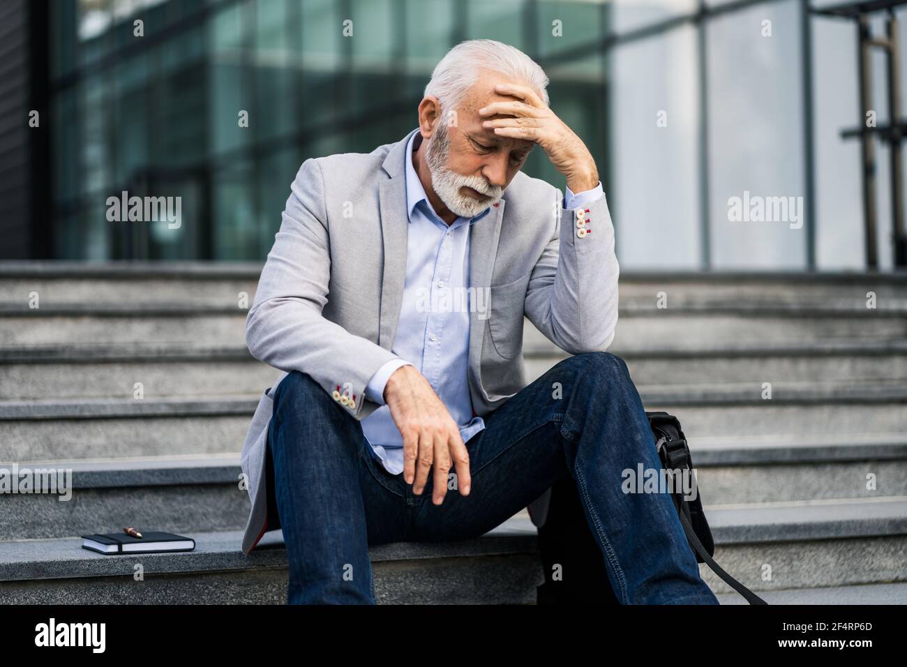 L'uomo d'affari anziano è stanco di lavoro. È seduto di fronte alla costruzione dell'azienda e ha mal di testa. Foto Stock