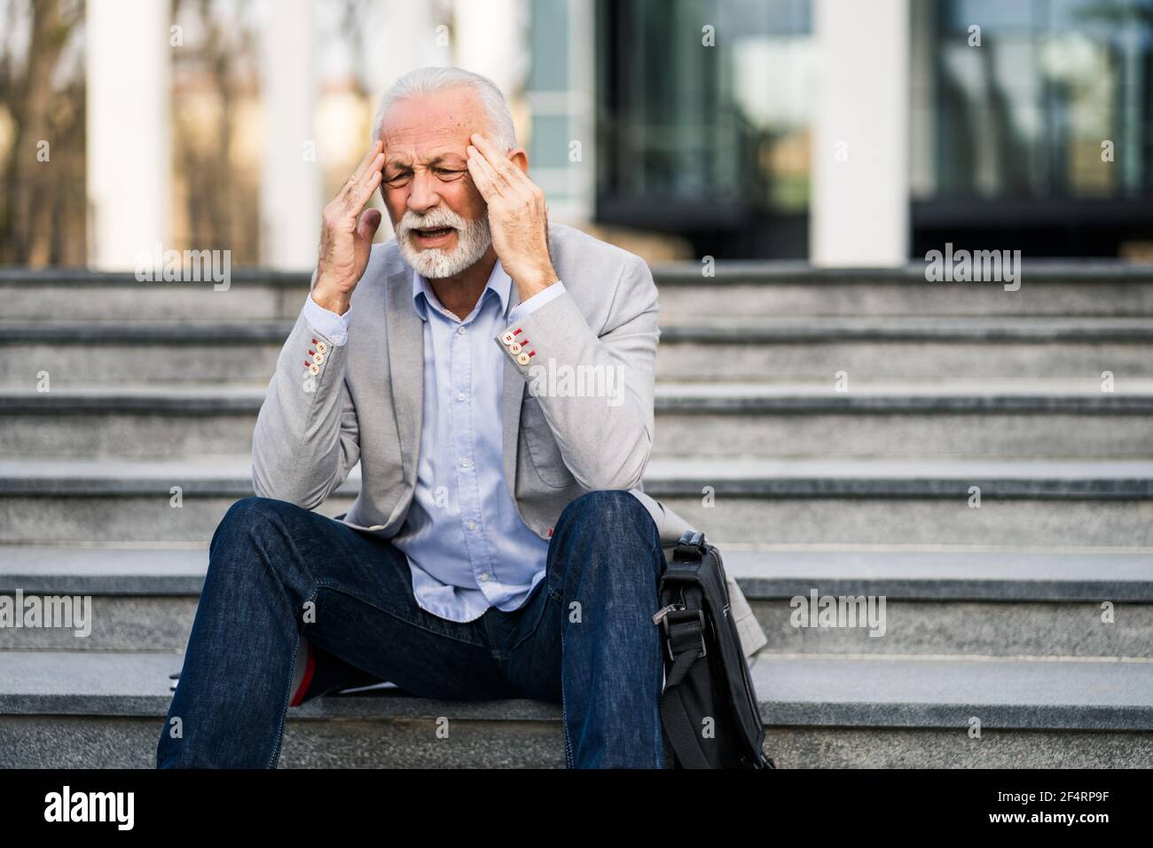 L'uomo d'affari anziano è stanco di lavoro. È seduto di fronte alla costruzione dell'azienda e ha mal di testa. Foto Stock