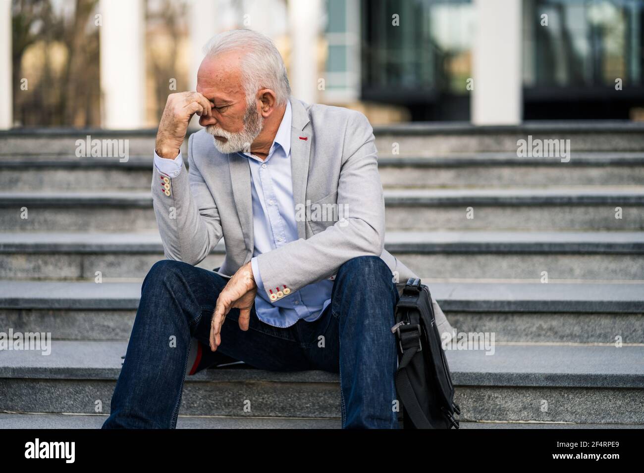 L'uomo d'affari anziano è stanco di lavoro. È seduto di fronte alla costruzione dell'azienda e ha mal di testa. Foto Stock