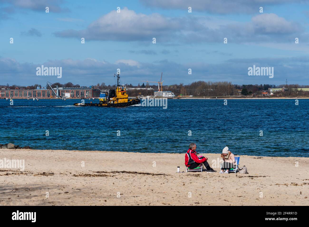 Stand an der Kieler Förde im Frühjahr, ein Paar hat es sich am Wasser gemütlich gemacht, ein Hafenschlepper passiert die Scene Foto Stock
