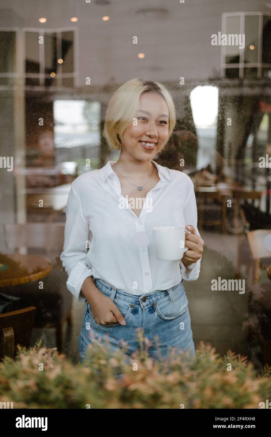 Giovane donna asiatica capelli corti con tazza di caffè è in piedi al coperto dietro una grande finestra, guardando fuori sorridente. Foto Stock