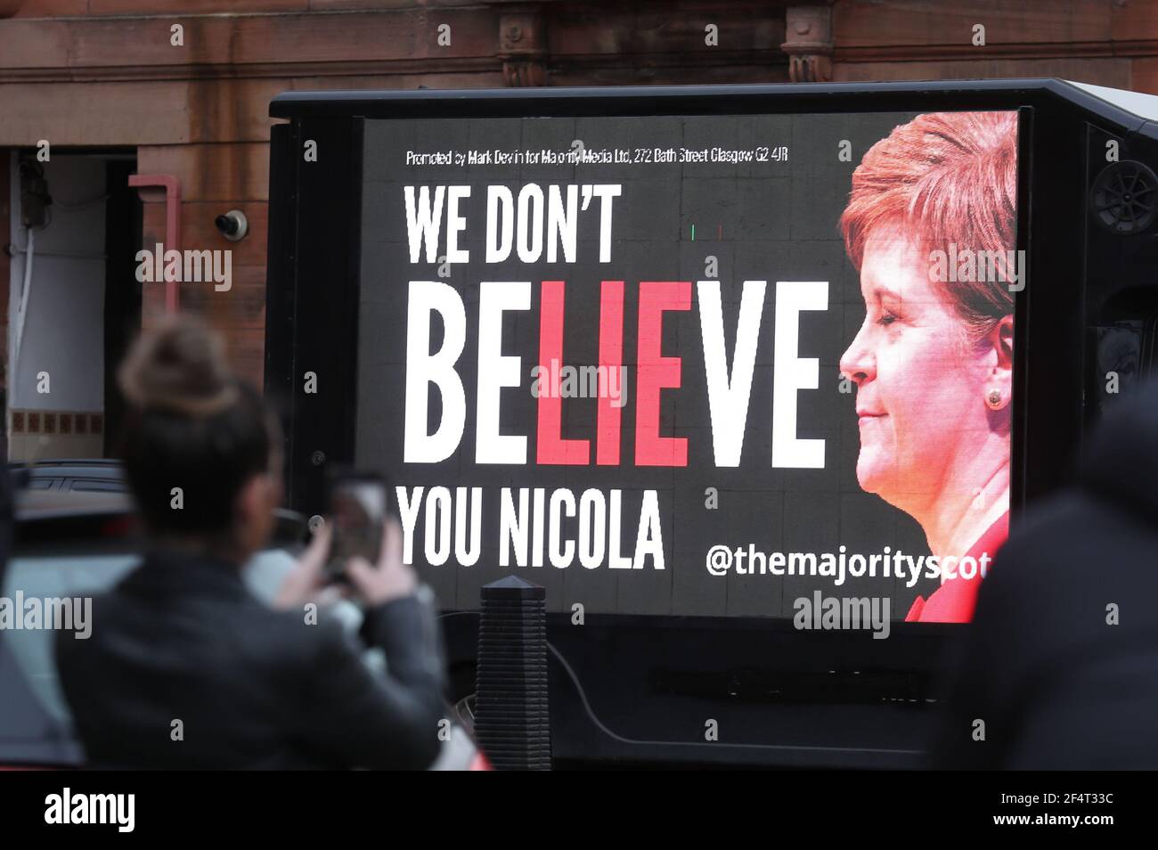 Una donna scatta una foto di un tabellone mobile che dice "non ci crediamo Nicola" mentre guida attraverso la circoscrizione del primo ministro Nicola Sturgeon nella zona di Govanhill a Glasgow come parte della campagna di ResignSturgeon della maggioranza nella corsa alle elezioni scozzesi del 6 maggio. Data immagine: Martedì 23 marzo 2021. Foto Stock