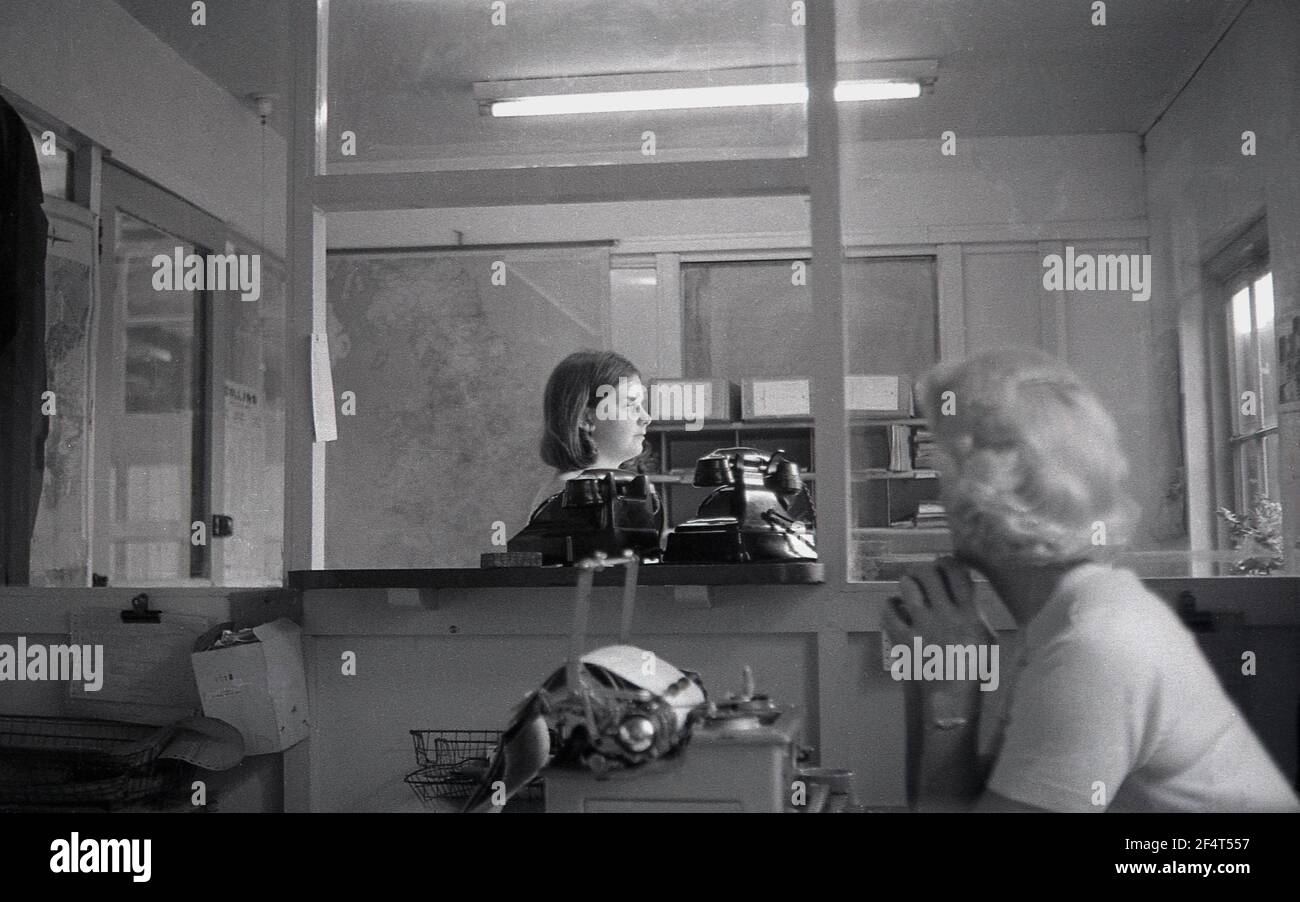 1964, storico, daydreaming.... all'interno dei locali di un'azienda industriale, una donna matura in un ufficio parted seduto ad una macchina da scrivere che guarda attraverso la finestra partizionata ad un'altra, donna più giovane nell'ufficio accanto in piedi da due grandi vecchi telefoni bakerlite dell'era che guarda fuori, Inghilterra, Regno Unito. Foto Stock