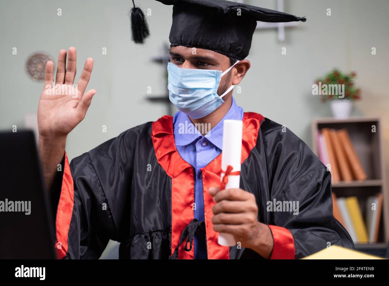 Studente con maschera medica che partecipa a una cerimonia virtuale di laurea video da casa su laptop: concetto di assistenza sanitaria, sicurezza e novità normale Foto Stock