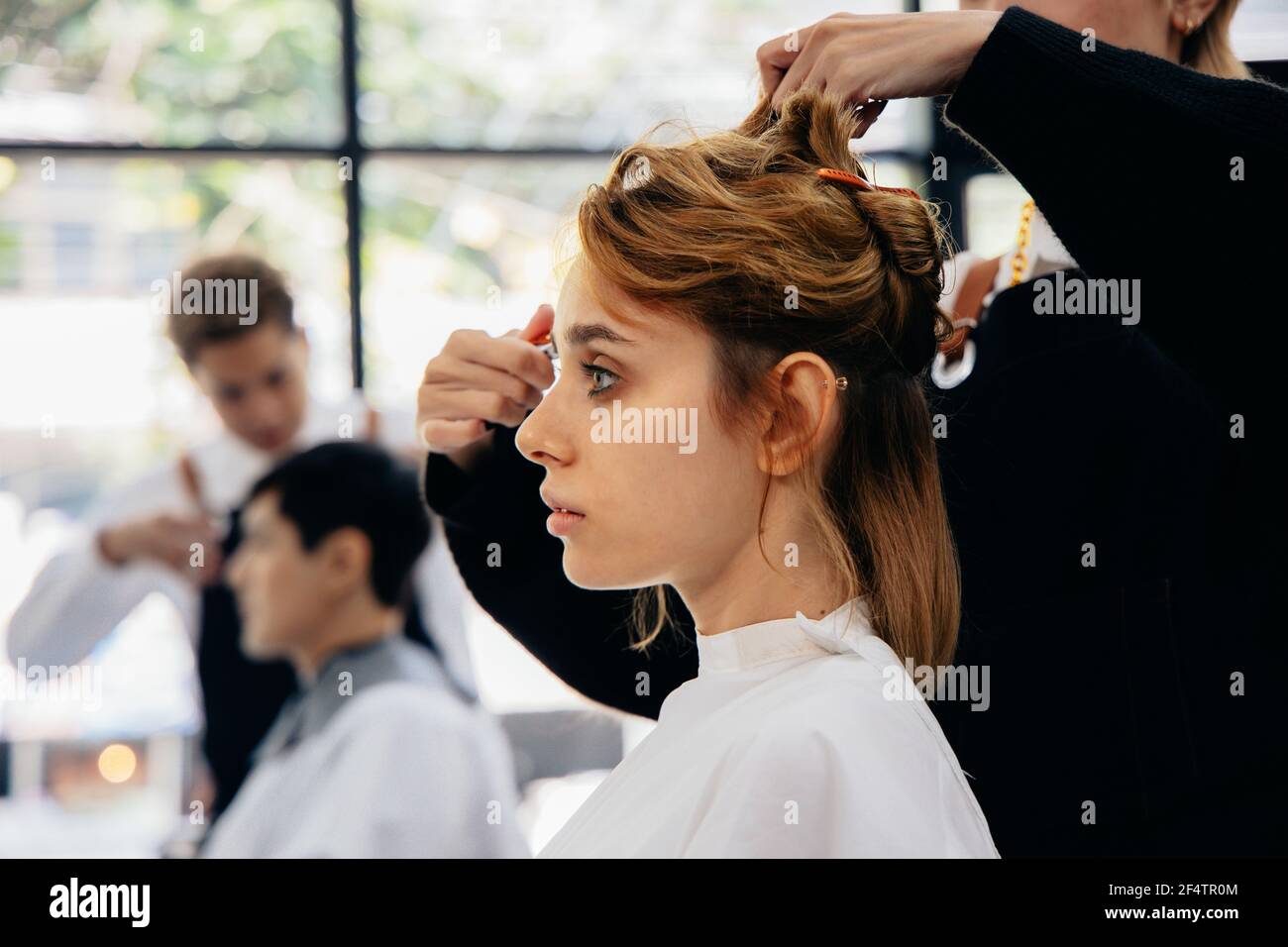 Bella giovane caucasica seduta in salotto moderno con avvolgente intorno ad ottenere un taglio di capelli con acconciatura da parte del parrucchiere femminile che indossa grembiule Foto Stock