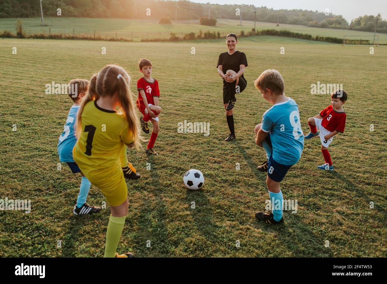 I bambini si allenano con il loro allenatore all'esterno. Gruppo di bambini che si allungano insieme con la loro allenatrice femminile prima di giocare a calcio in campo al tramonto. Foto Stock