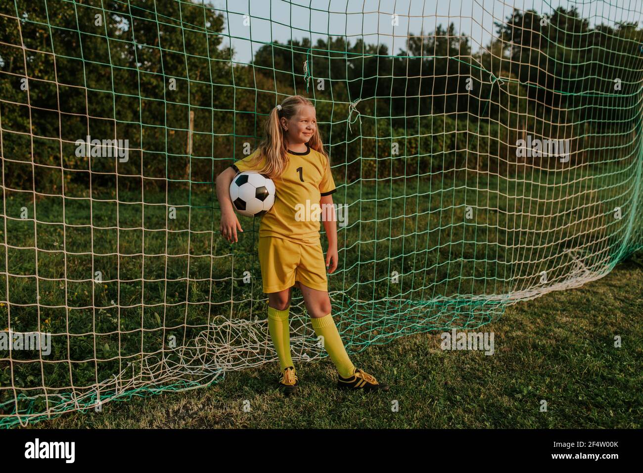 Portiere ragazza con palla in piedi al gol sul campo di calcio. Intera lunghezza di ragazza in abito da calcio giallo davanti alla rete di calcio. Foto Stock