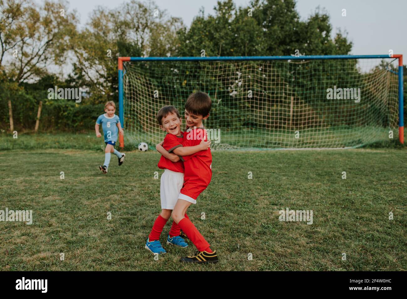 Bambini che giocano a calcio all'aperto sul campo. Lunghezza completa dei ragazzi felici che abbracciano dopo aver segnato l'obiettivo durante la partita di calcio amatoriale. Foto Stock