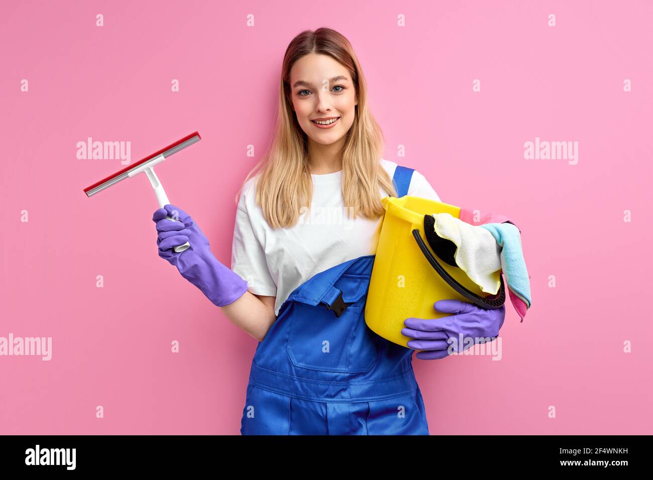 Sorridente bella donna caucasica in tute blu e guanti di gomma guardare fotocamera che tiene i materiali di pulizia. Isolato sfondo rosa, ritratto di prett Foto Stock