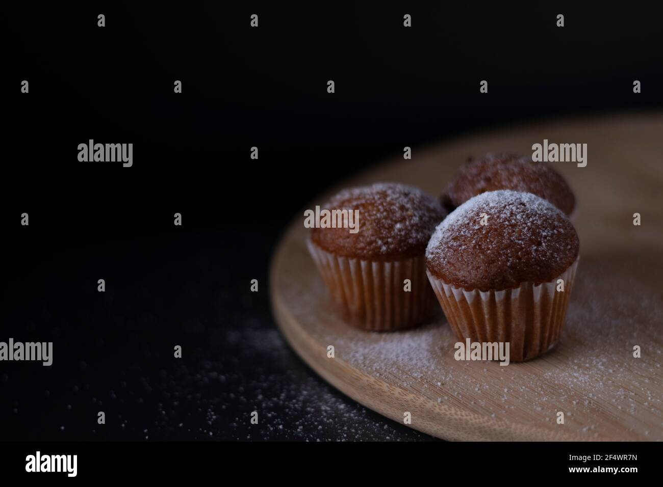 tre muffin ricoperti di zucchero in polvere su supporto di legno isolato su sfondo nero scuro. dolce da forno Foto Stock