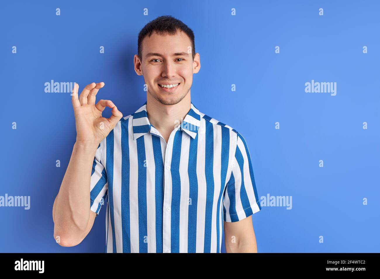 Bell'uomo che mostra un gesto e un sorriso OK, si sente felice, di buon umore. Ragazzo in camicia a righe blu che si posa isolato su sfondo blu, copia spazio Foto Stock
