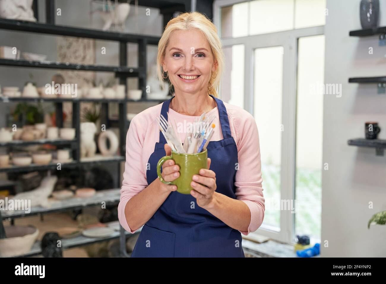 Pronto per iniziare. Ritratto di donna felice in grembiule blu che tiene gli attrezzi, spazzole per la creazione di ciotola di ceramica di argilla fatta a mano in laboratorio di ceramica Foto Stock