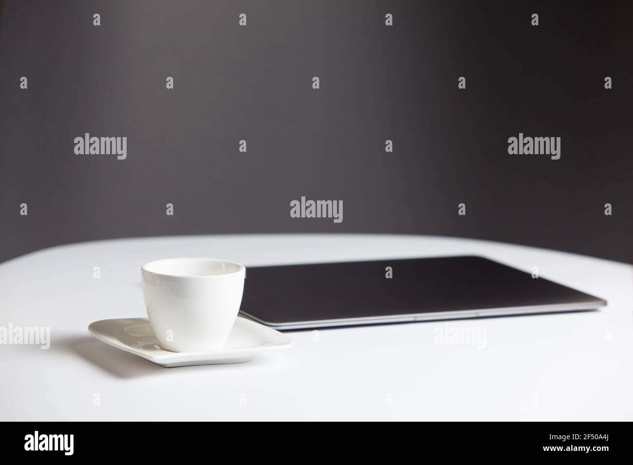 Composizione con un moderno computer portatile chiuso e una tazza di caffè con piattino sul tavolo bianco su sfondo grigio. Postazione di lavoro remota, ufficio domestico. Foto Stock