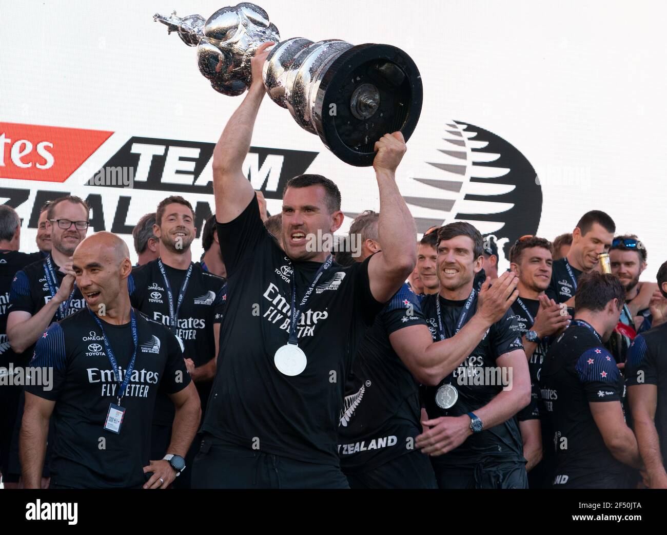 Auckland, Nuova Zelanda, 17 marzo 2021 - il macinacaffè neozelandese Joe Sullivan tiene in palio la America's Cup durante la cerimonia di presentazione dopo aver mantenuto la America's Cup dopo la sconfitta degli sfidanti italiani Luna Rossa Prada Pirelli. Dietro a destra è lo skipper Peter che scava con altri membri dell'equipaggio festeggiando. Credito: Rob Taggart/Alamy Foto Stock