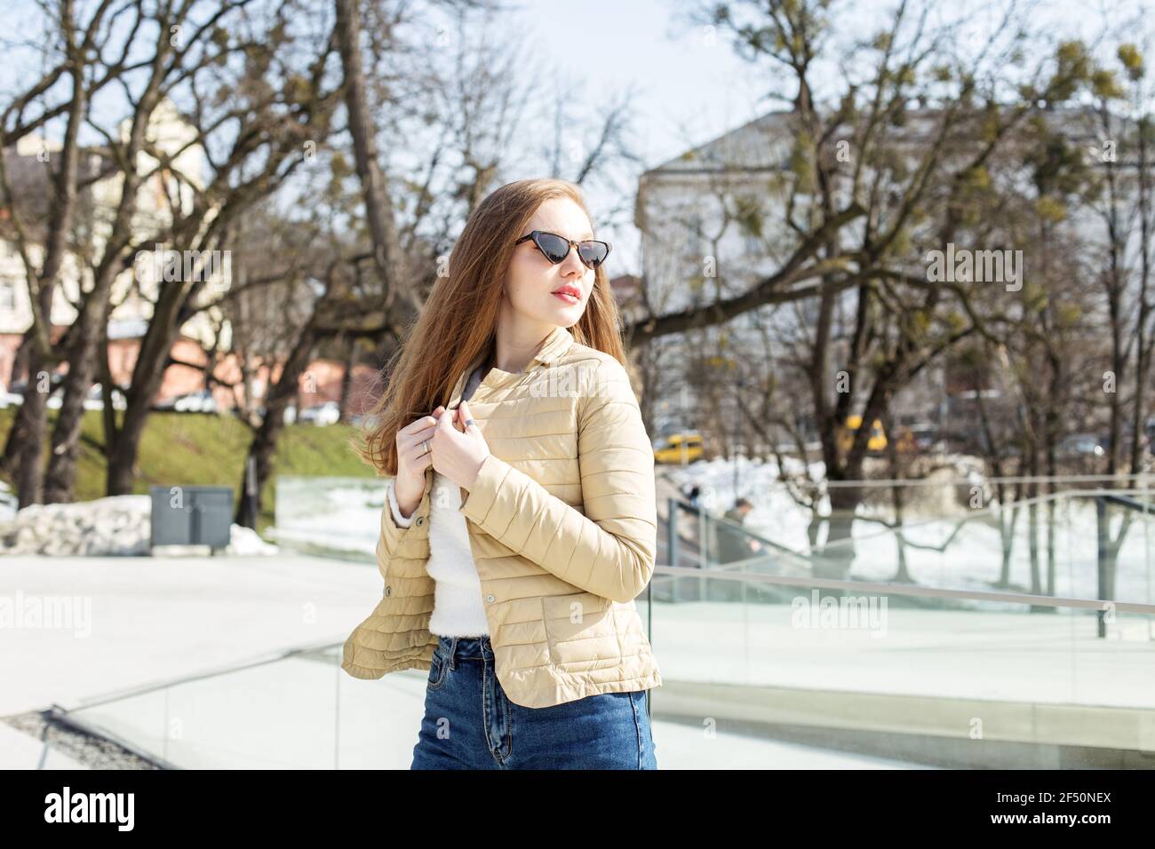 Una bella ragazza bionda in una breve giacca leggera e occhiali da sole cammina per la città, godendosi il tempo di primavera. Foto Stock