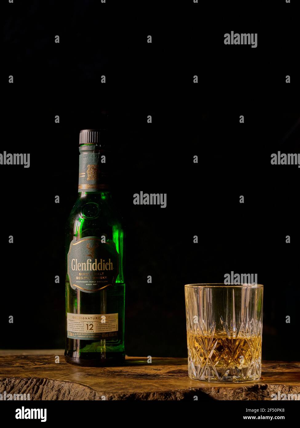 Glenfiddich Scotch Whisky bottiglia e vetro mezzo riempito su un asse di legno di oliva su uno sfondo nero scuro Foto Stock