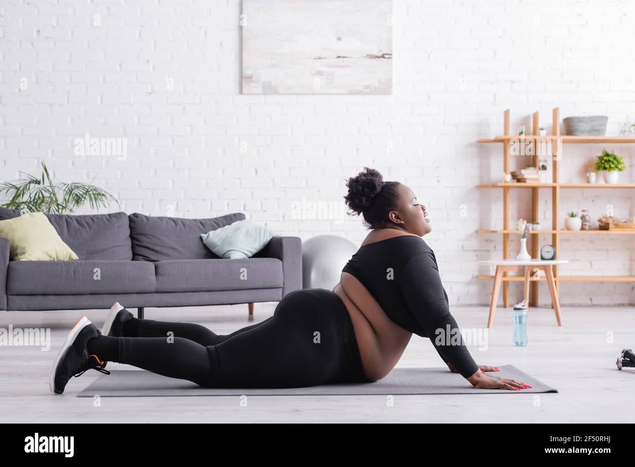 vista laterale della donna afroamericana plus size in abbigliamento sportivo esercitarsi sul tappetino di idoneità in soggiorno Foto Stock