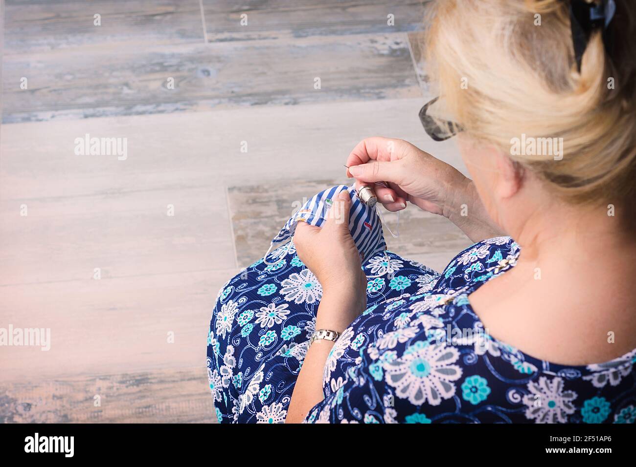 immagine da dietro di una donna bionda in un blu vestito con fiori stampati con i suoi capelli raccolti a mano cucire una maschera per bambini e pavimento rustico Foto Stock