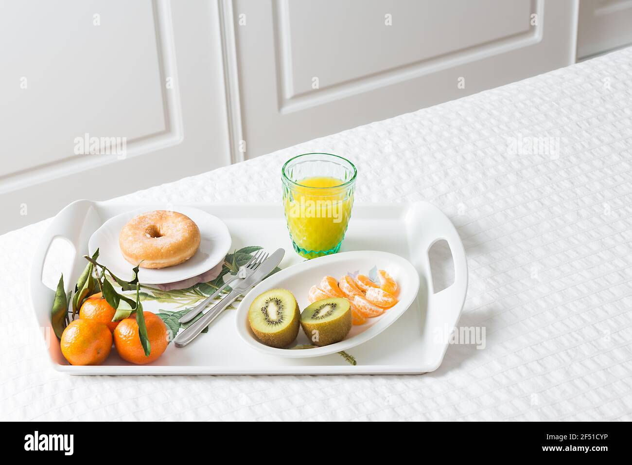 Immagine orizzontale di un vassoio con una ciambella per la colazione kiwi mandarino e succo d'arancia su un letto bianco con porte di fondo bianche Foto Stock