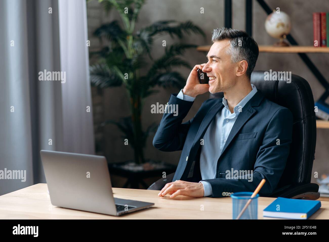 Riuscito agente immobiliare adulto caucasico sicuro di sé, seduto in ufficio moderno, in abito elegante, parlando su un cellulare con un cliente, guardando via, sorridente amichevole Foto Stock
