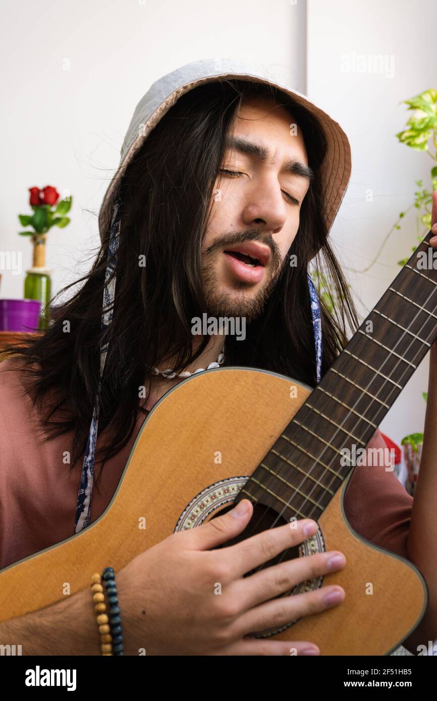 giovane uomo dai capelli lunghi che suona la chitarra acustica a casa e  canta. giovane bohémien. indossa una camicia rosa con un cappello a  secchio. immagine verticale Foto stock - Alamy