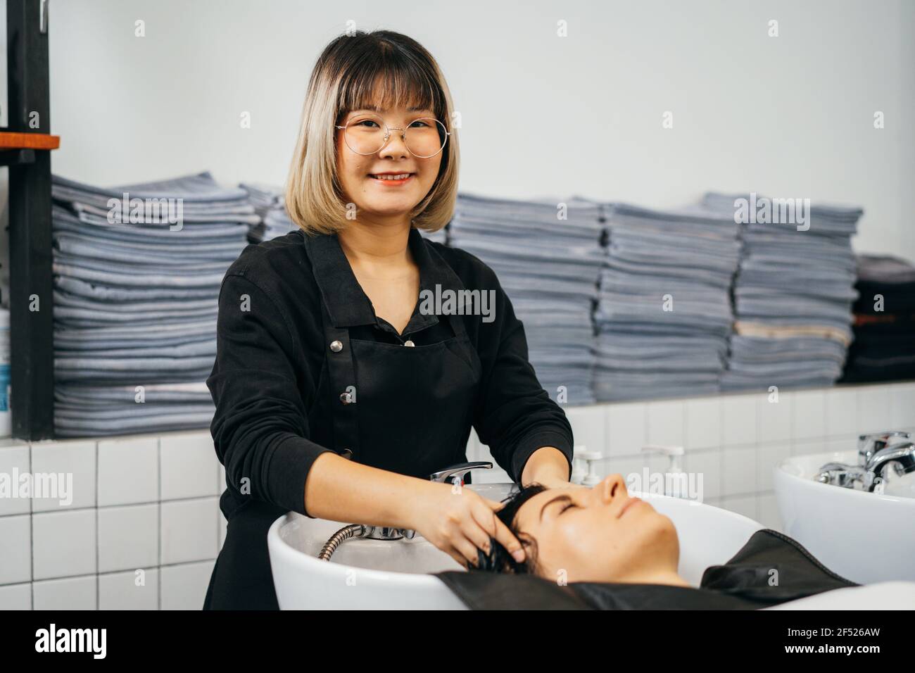 Ritratto di giovane estetista asiatica guardando la macchina fotografica, in piedi e massaggiando i capelli e la testa di giovane cliente femminile con la testa in lavabo sorridente e guardando la macchina fotografica Foto Stock
