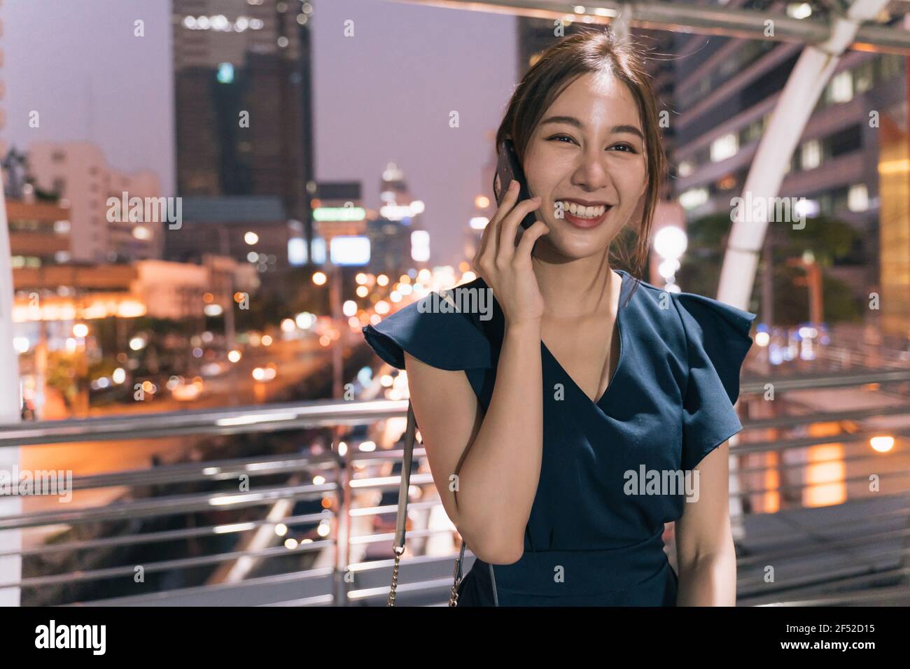Ritratto di sorridente giovane asiatica riuscita donna in abbigliamento casual parlare sullo smartphone mentre ci si trova sul ponte pedonale con vista sulla città in background Foto Stock