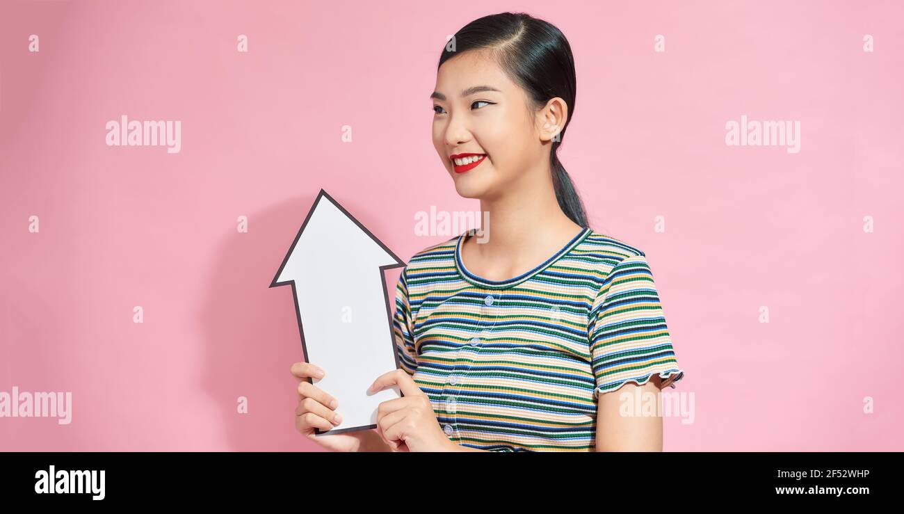 felice sorridente giovane donna asiatica che tiene grande bianca freccia spessa puntando su sfondo rosa Foto Stock