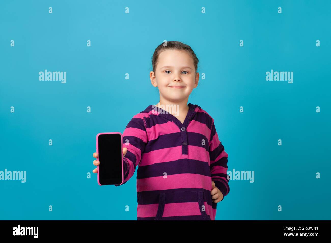 il ritratto in primo piano di una ragazza felice, sorridente e positiva in una felpa con cappuccio a righe viola mostra lo schermo del telefono in una custodia rosa, isolata su uno sfondo blu Foto Stock