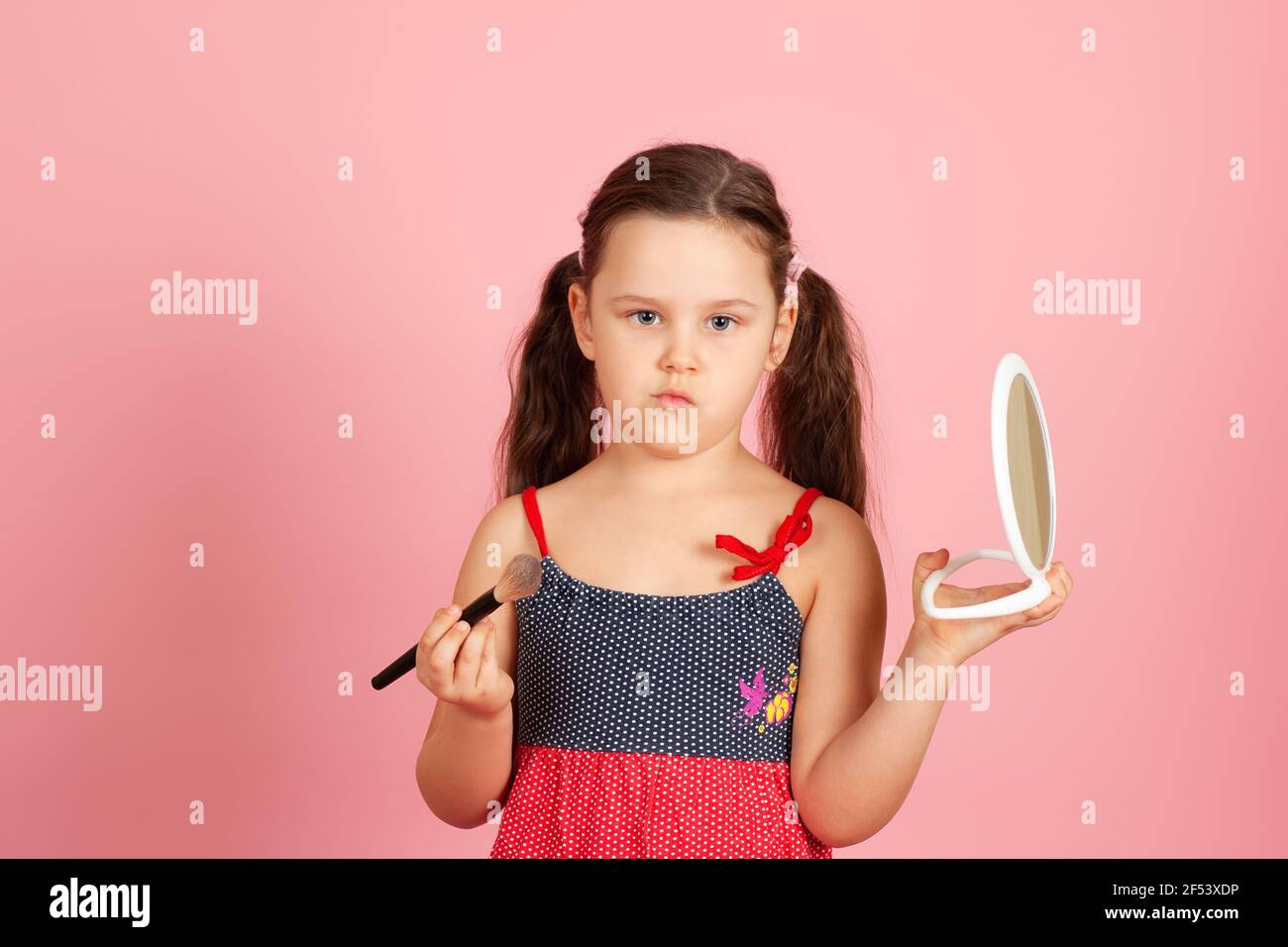 una ragazza pensiva con coda di pony tiene un pennello di polvere e uno specchio bianco di plastica, facendo un arco di spugna, isolato su uno sfondo rosa Foto Stock