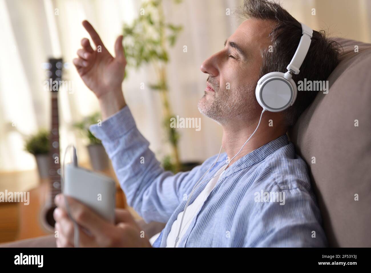 Dettaglio dell'uomo seduto sul divano a casa ascoltando alla musica proveniente da un telefono cellulare con cuffie bianche entrare in una luce solare calda attraverso il th Foto Stock