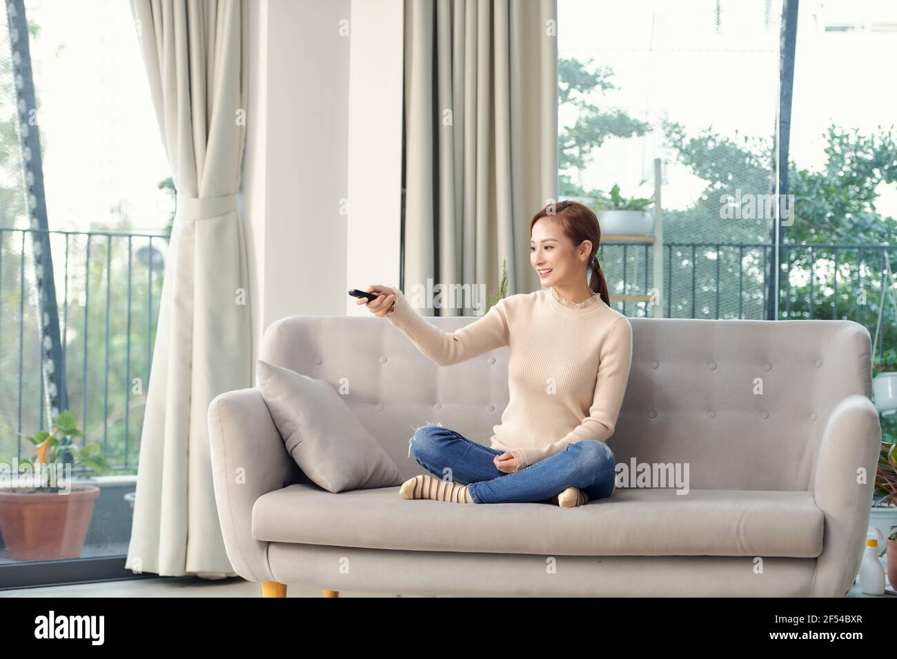 Immagine di una donna giovane ottimista e sorridente positiva seduto in casa guardare la tv tenendo il telecomando sul divano. Foto Stock