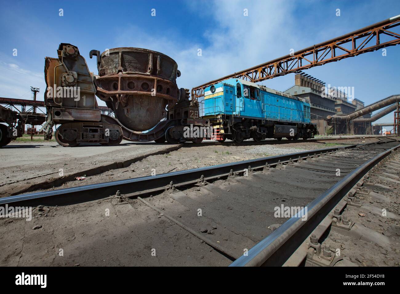 Impianto di leghe metalliche. Locomotiva blu e carri auto scorie arrugginite. Impianto metallurgico edificio industriale principale sullo sfondo. Cielo blu. Foto Stock