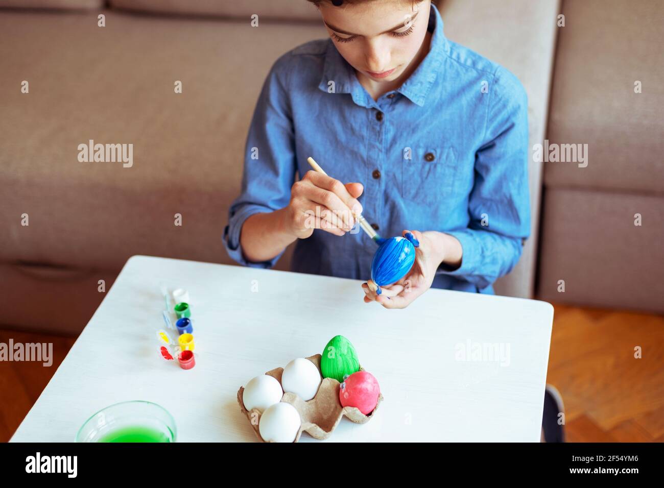Bella ragazza di nove anni in camicia blu dipinge le uova ad un tavolo bianco. Concetto di Pasqua. Messa a fuoco selettiva. Foto Stock
