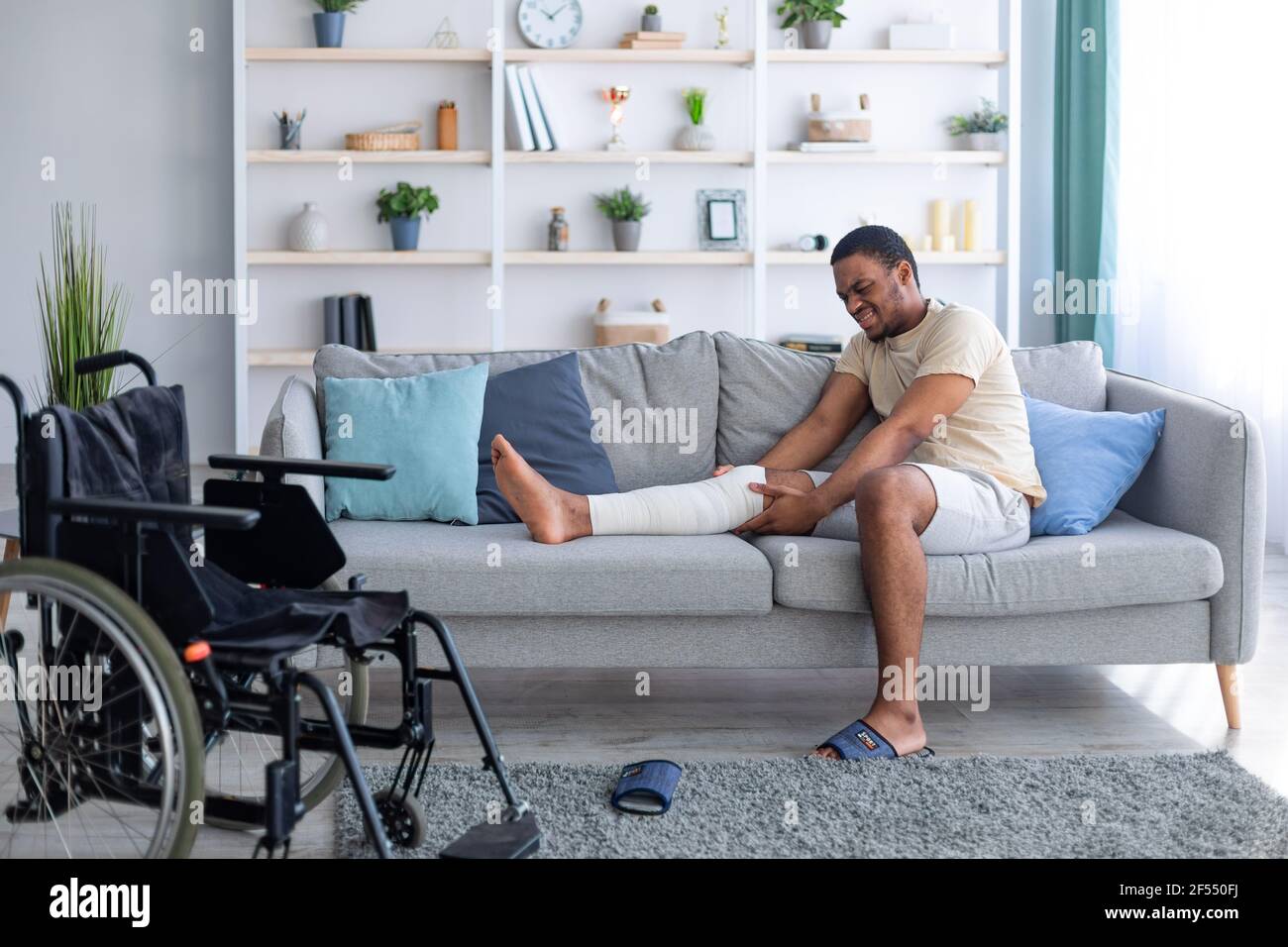 Uomo nero millenario con gamba rotta intonacata, che soffre di dolore sul divano a casa, sedia a rotelle in piedi nelle vicinanze Foto Stock