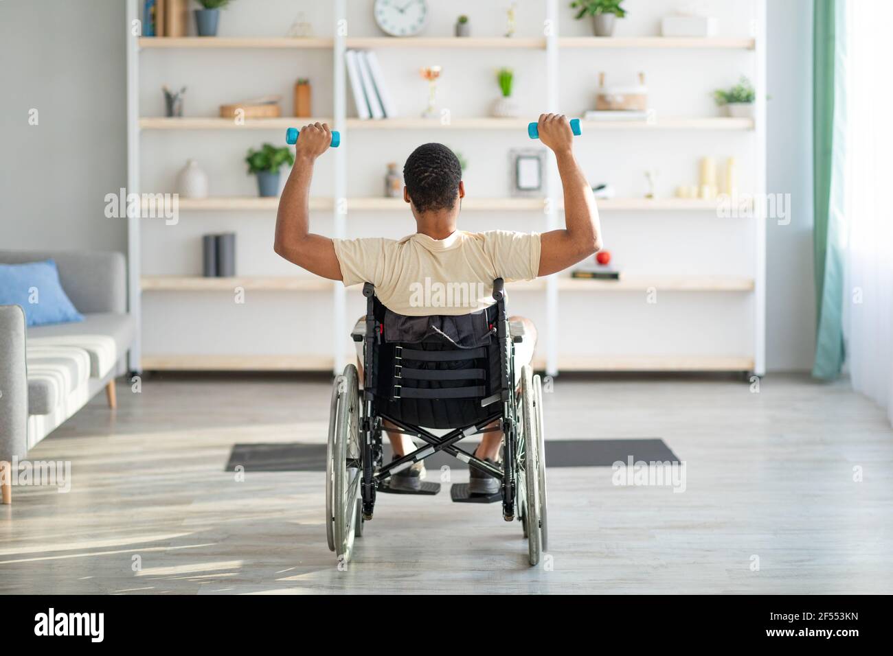 Vista posteriore di un ragazzo nero con disabilità in sedia a rotelle che si esercita con manubri a casa. Concetto di sport e disabilità Foto Stock