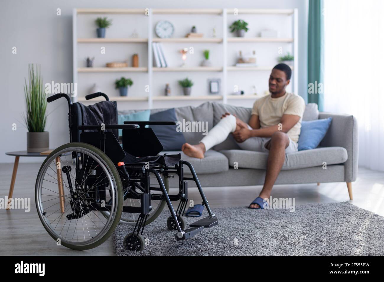 Giovane ragazzo nero con gamba rotta intonacata seduta sul divano, messa a fuoco su sedia a rotelle vuota, spazio di copia Foto Stock