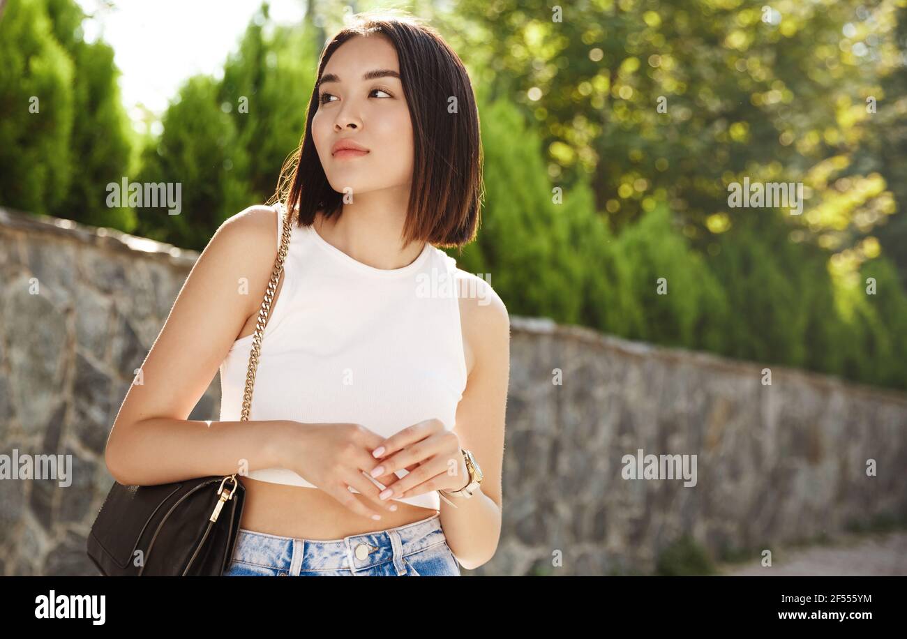 Elegante donna asiatica in attesa di qualcuno nel parco. Ragazza moderna in abiti estivi guardando a sinistra, avendo una data o riunione in città Foto Stock