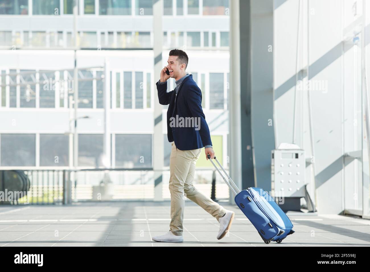 Ritratto laterale a lunghezza intera di uomo d'affari informale che cammina dentro stazione con telefono e valigia Foto Stock