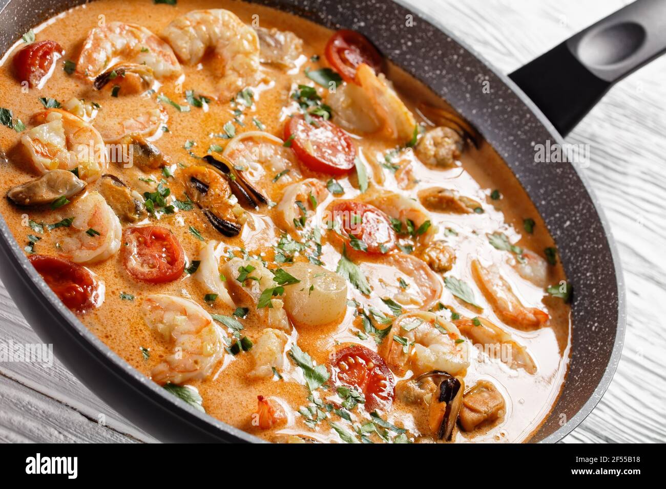 Pesce saltato con gamberi, fette di pesce bianco, capesante, pomodori ciliegini in una padella su un tavolo di legno, primo piano, cucina francese Foto Stock