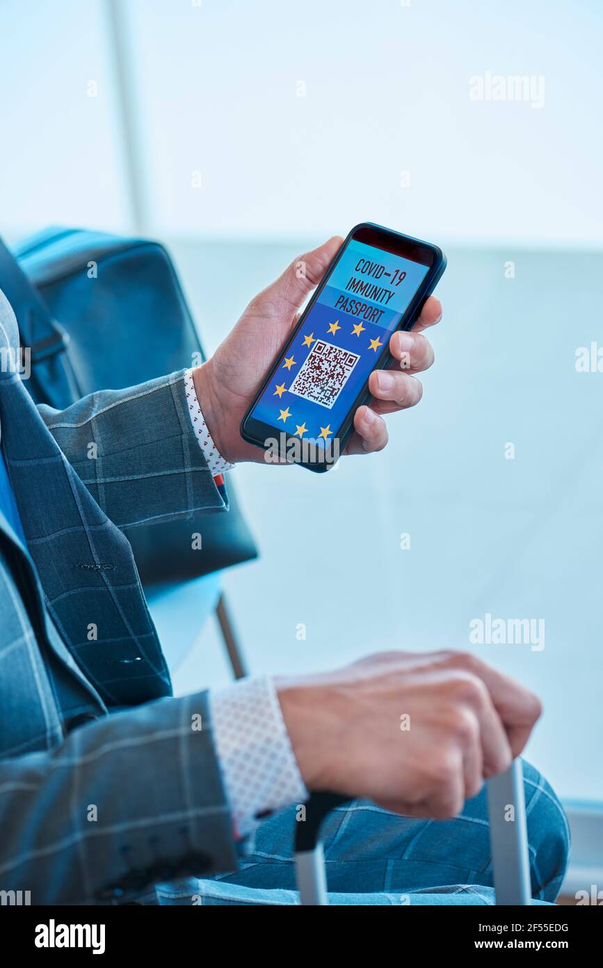 un giovane uomo d'affari caucasico, che indossa una tuta grigia, seduto nella sala d'attesa di un aeroporto, ha una simulazione elettronica europea covid-19 immunità p Foto Stock